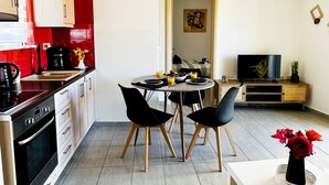A modern kitchen with a red backsplash, a dining area with two black chairs, a TV on a wooden stand, and a small plant on the side table. - Cozy Snuggle- 2min To Beach, 6min Walk To Old Town (Chania)