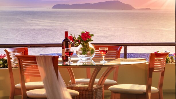 Una mesa con sillas, una botella de vino y un florero con flores, con vista a un cuerpo de agua y tierra a lo lejos.