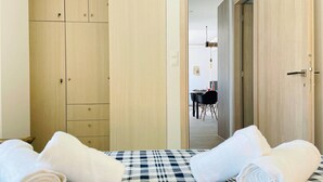 A room with a bed, a wooden wardrobe, and a dining area visible through an open door.