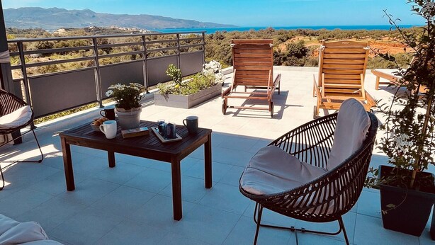A terrace with two wooden chairs, a table with a potted plant, and a view of the sea and hills.