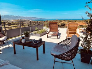 A terrace with two wooden chairs, a table with a potted plant, and a view of the sea and hills.
