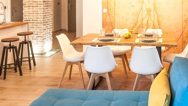A modern living room with a blue sofa, a dining area with a wooden table, and a large wall mural. - Secret Loft, 2min Walk From The Beach, High Tech (Chania)