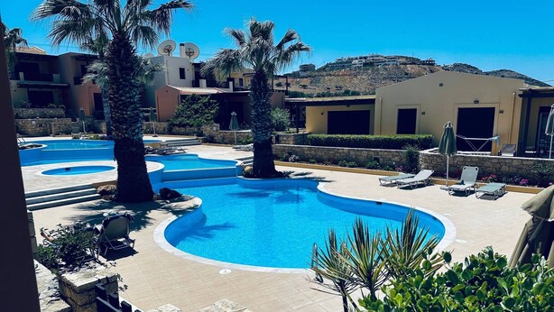 Una piscina con varias secciones, rodeada de palmeras y sillas de descanso, con edificios al fondo.