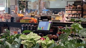 Interieur van een supermarkt met een zelfservicekassa, diverse groenten en fruit, en potplanten.