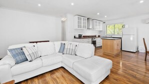 Een moderne woonkamer met een witte hoekbank, houten vloeren en een keuken met witte kastjes en een koelkast.