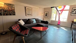 A modern living room with a sofa, a red coffee table, and a plaid chair. There are framed artworks on the walls and a window with curtains.