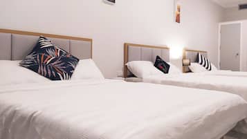 Habitación de hotel con dos camas individuales, cada una con cabecero de madera, ropa de cama blanca y almohadas decorativas. Hay una mesita de noche pequeña con una lámpara y un cuadro enmarcado en la pared sobre las camas.