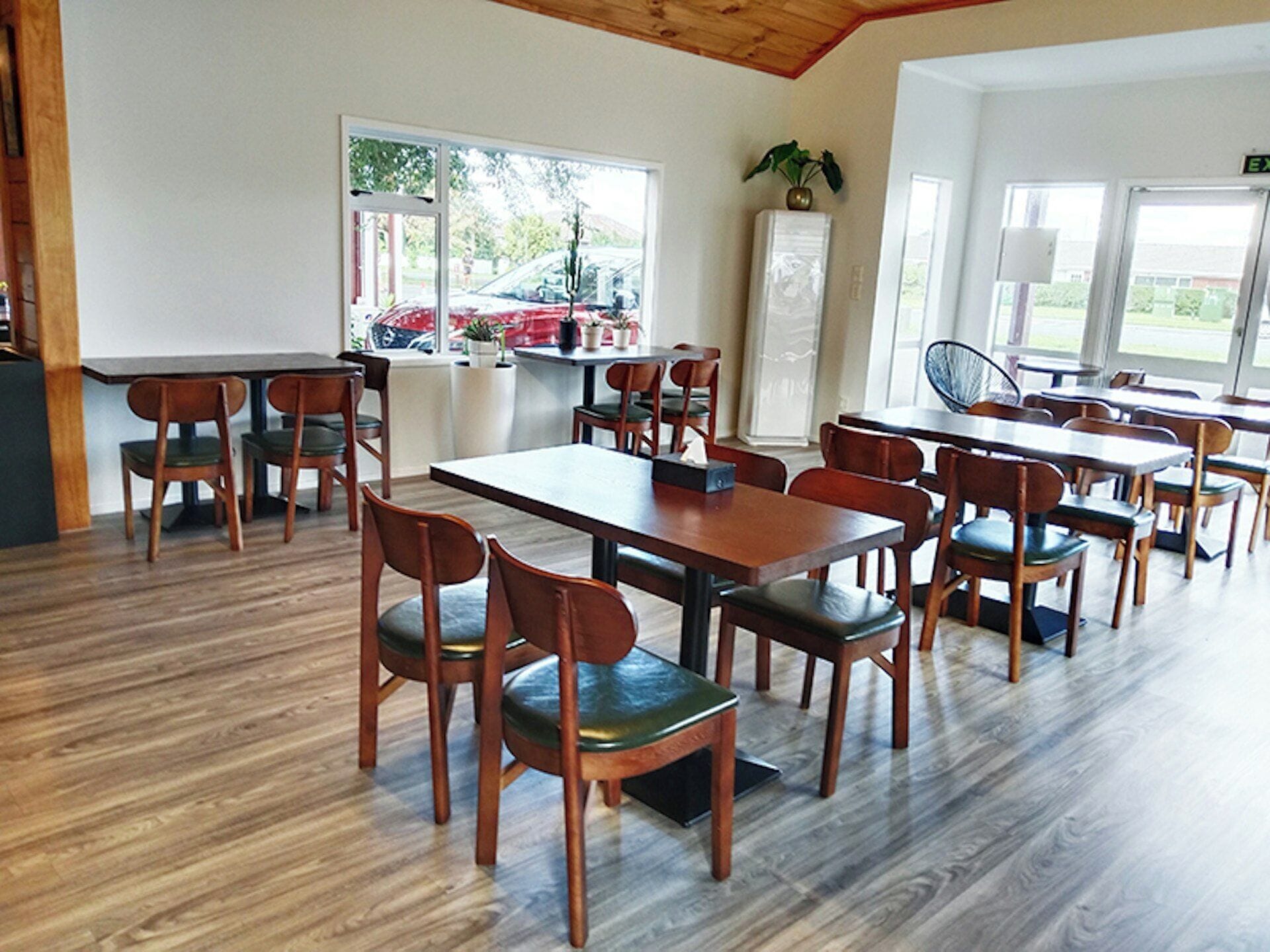 Un comedor con mesas y sillas de madera, ventanas amplias con vistas al exterior y una planta en maceta sobre un mueble.