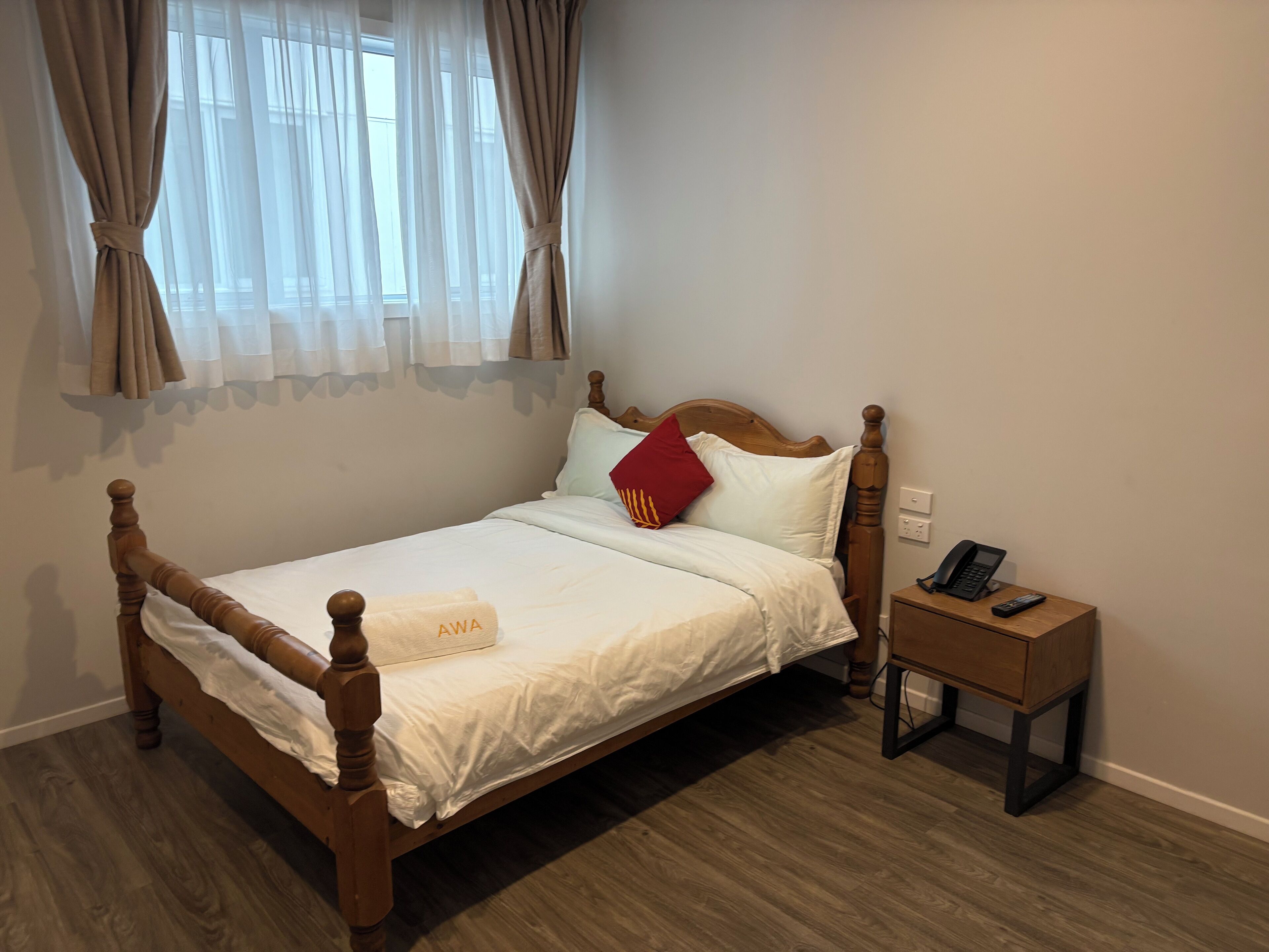 A single bed with a wooden frame, white bedding, and a red pillow. There is a bedside table with a phone and remote control.