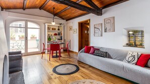 Een moderne woonkamer met een houten plafond, een grijze bank met rode en zwarte kussens, een eethoek met rode stoelen en een boekenkast.
