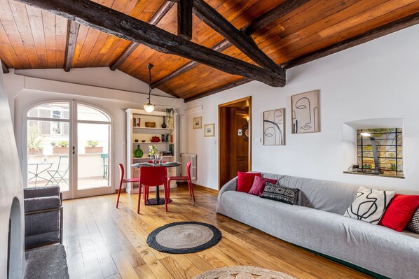 Een moderne woonkamer met een houten plafond, een grijze bank met rode en zwarte kussens, een eethoek met rode stoelen en een boekenkast.
