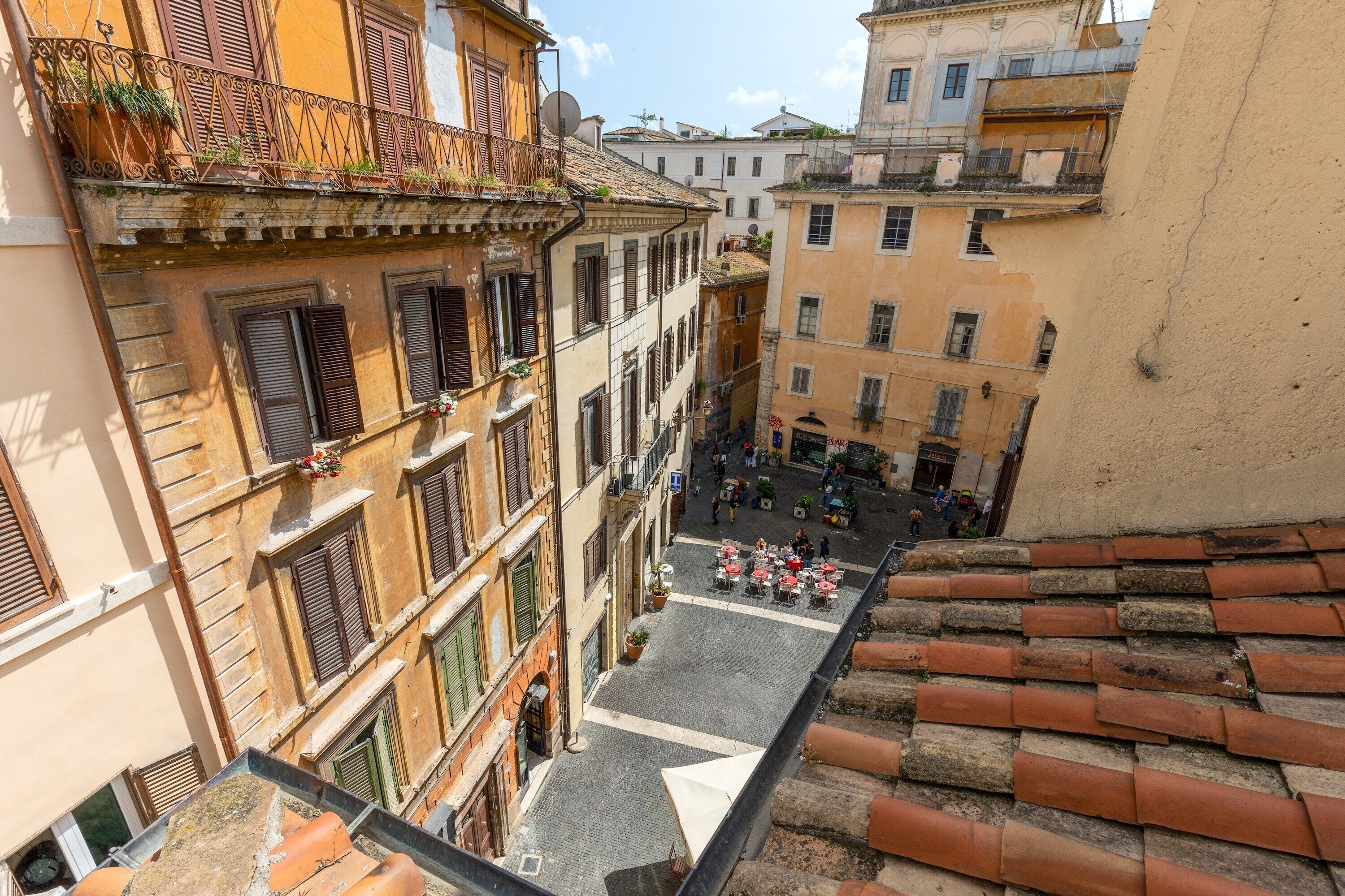 A narrow street with buildings on either side, featuring balconies with plants and a cobblestone street.