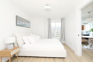 A modern bedroom with a large bed, a bedside table with a lamp, a view of the city through a window, and a sign indicating the room is a beach house. - 2 Bed + DEN 2 Bath Downtown Kelowna (Kelowna)