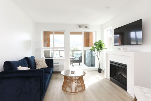 A modern living room with a dark blue sofa, a glass coffee table, a fireplace, and a large TV.