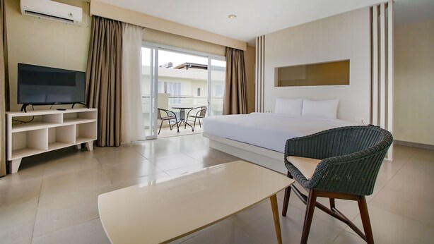 A hotel room with a bed, a TV on a stand, a wicker chair, a small table, and a balcony with a view of buildings.