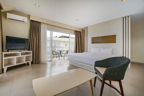 A hotel room with a bed, a TV on a stand, a wicker chair, a small table, and a balcony with a view of buildings.