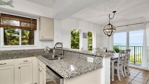 Una cocina con encimeras de granito, armarios blancos, lavavajillas integrado y un comedor con vista al océano.