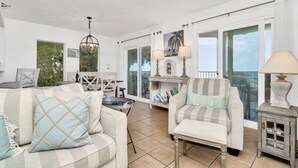 Amplia sala de estar con sofá, sillón y una pequeña mesa. Hay dos lámparas, una araña y vistas al exterior a través de ventanas grandes.