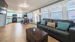 A spacious living room with a dark leather sofa, a coffee table with a plant, and a kitchen area with teal cabinets and modern appliances.