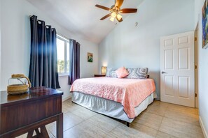 A bedroom with a bed, a ceiling fan, a window with curtains, a bedside table with a lamp, and a door.