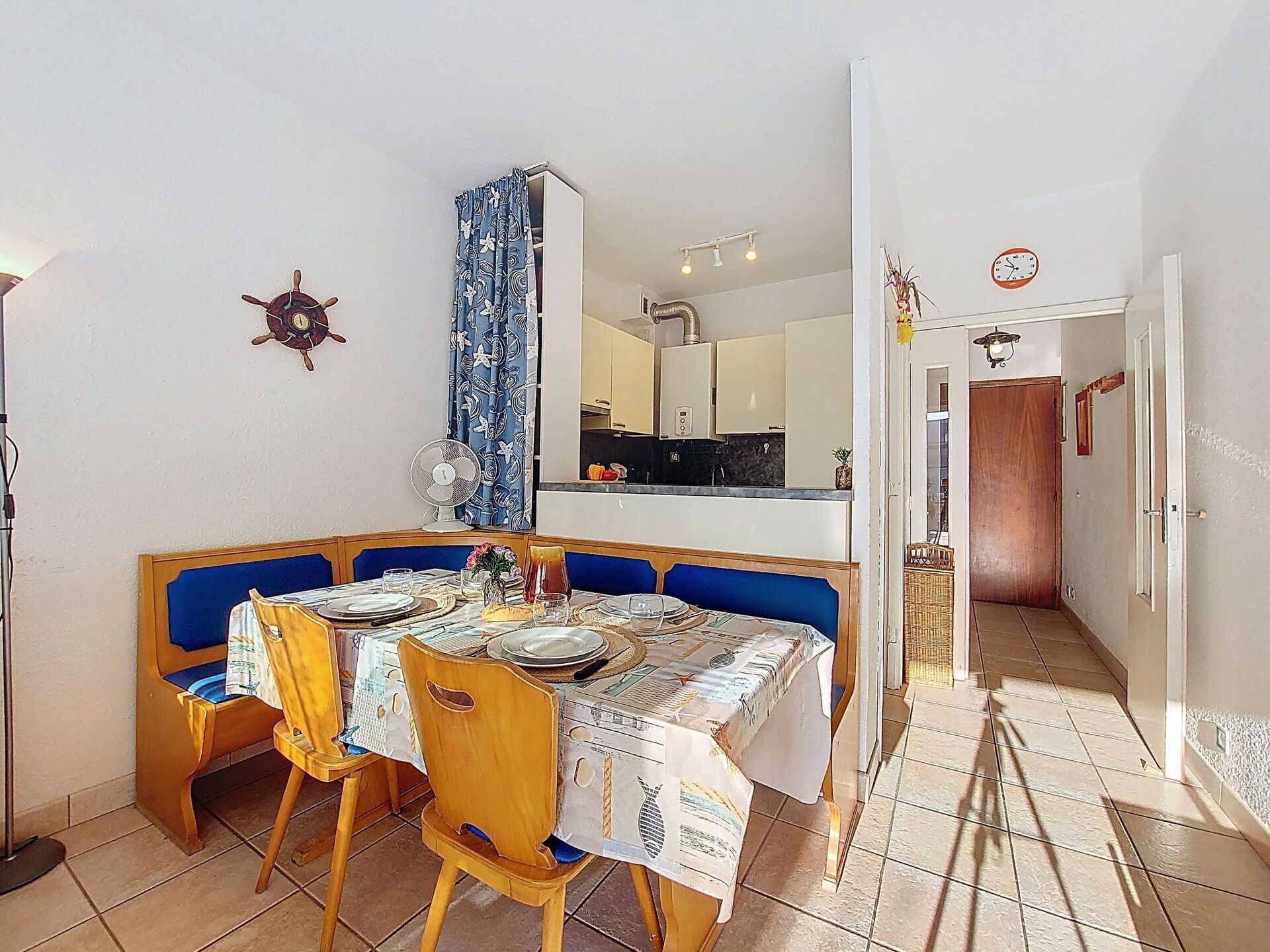 A dining area with a table set for four, a blue upholstered bench, and a fan on the wall.