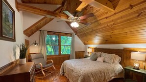 Une chambre confortable avec un plafond en bois, un lit recouvert d’une courtepointe à motifs, un fauteuil à bascule, un bureau avec des tiroirs, une fenêtre avec des rideaux et un ventilateur de plafond.