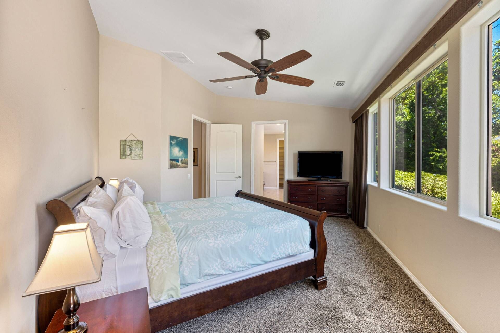 A bedroom with a bed, a ceiling fan, a TV, and a large window.