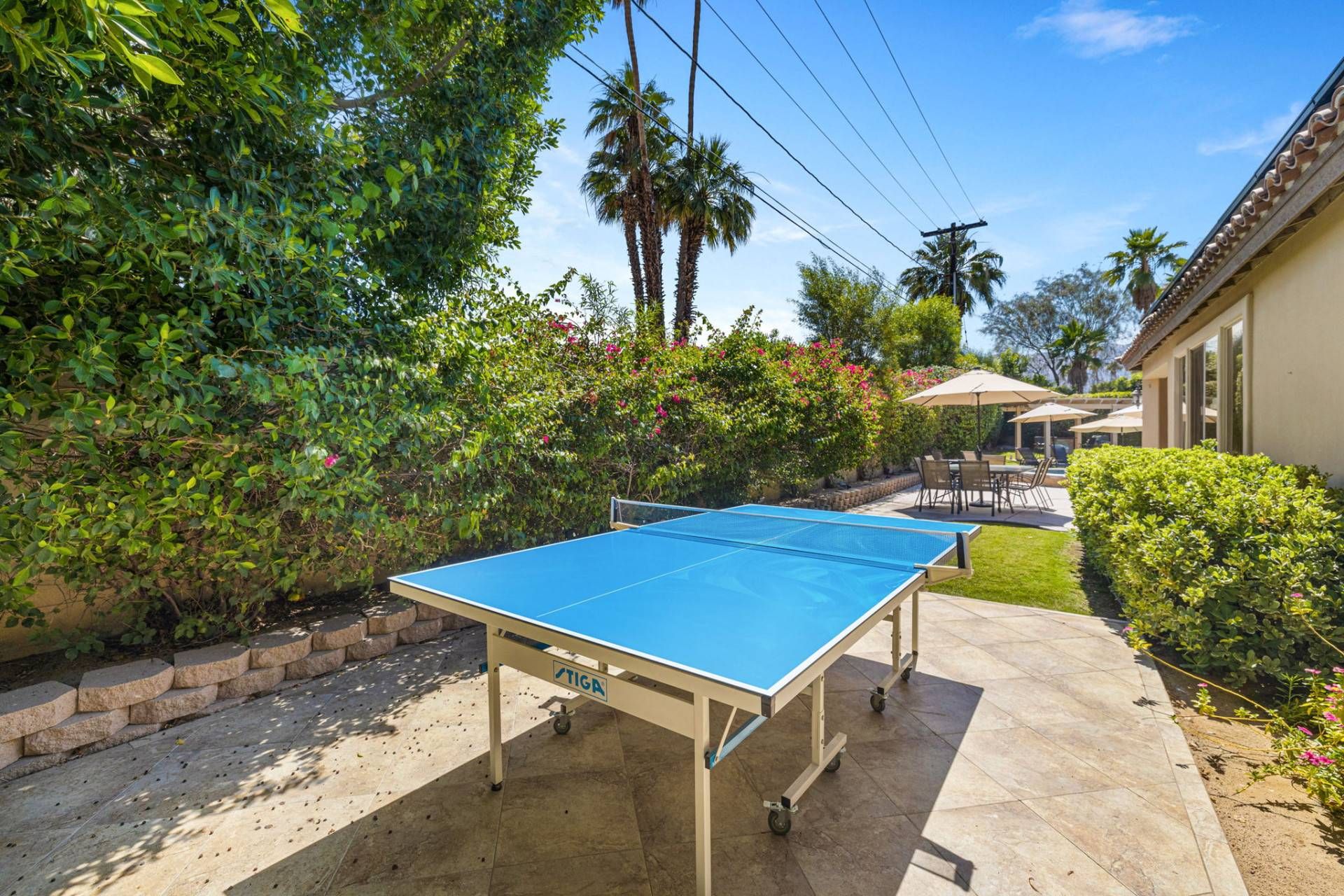 An outdoor ping pong table in a backyard with a patio area, surrounded by greenery and a house in the background.