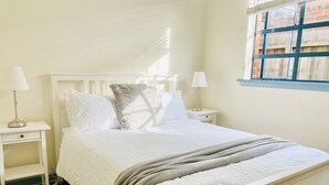 A neatly made bed with white bedding and a grey throw blanket, flanked by white nightstands with lamps, in a room with a window and wooden flooring. - Bay Rd | Kids & Pet Friendly | Free Parking (Cheltenham)