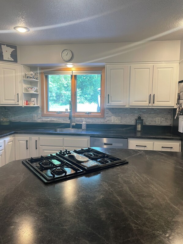 A kitchen with white cabinets, a stainless steel stove, a window with a view of greenery, and a clock on the wall.