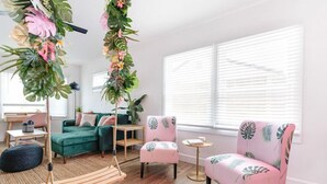 Un salon moderne avec un canapé vert, deux fauteuils roses ornés de motifs de feuilles, une plante suspendue, une balançoire en bois et une grande fenêtre avec des stores.
