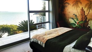 A bedroom with a large window, a bed with a striped blanket, a hanging light fixture, and a mural of palm trees. - Beachfront Luxury  (Clarks Beach)