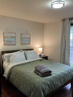 A neatly made bed with a green patterned bedspread, two floral pillows, and a folded towel on the bed. There are two framed paintings on the wall above the bed, a bedside lamp on a nightstand, and a window with curtains.
