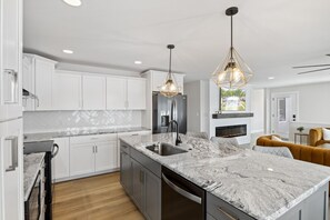 A modern kitchen with white cabinets, a granite countertop, and a built-in dishwasher. There are two pendant lights and a fireplace.