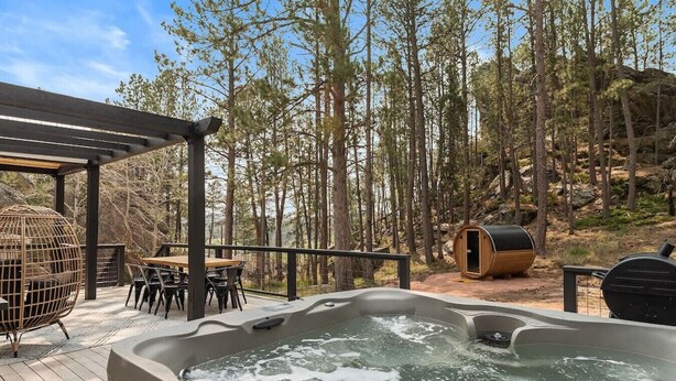 A hot tub surrounded by a wooden deck, a wooden table with chairs, and a wooden swing chair. - Little Italy- Hilltop Cottage (Custer)