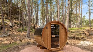 A wooden barrel-shaped structure with a door, situated in a forested area with a gravel path and a welcome mat. - Little Italy- Hilltop Cottage (Custer)