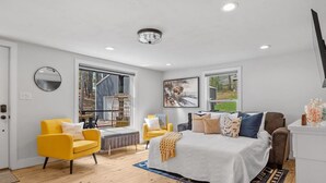 A bedroom with a large bed, two yellow chairs, a wooden floor, and a view of the outdoors. - Little Italy- Hilltop Cottage (Custer)