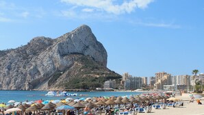 A beach with umbrellas and lounge chairs, a large rock formation, and a cityscape in the background. - Alexia II 123 - Grupo Turis - Calpe, Costa Blanca (Calpe)
