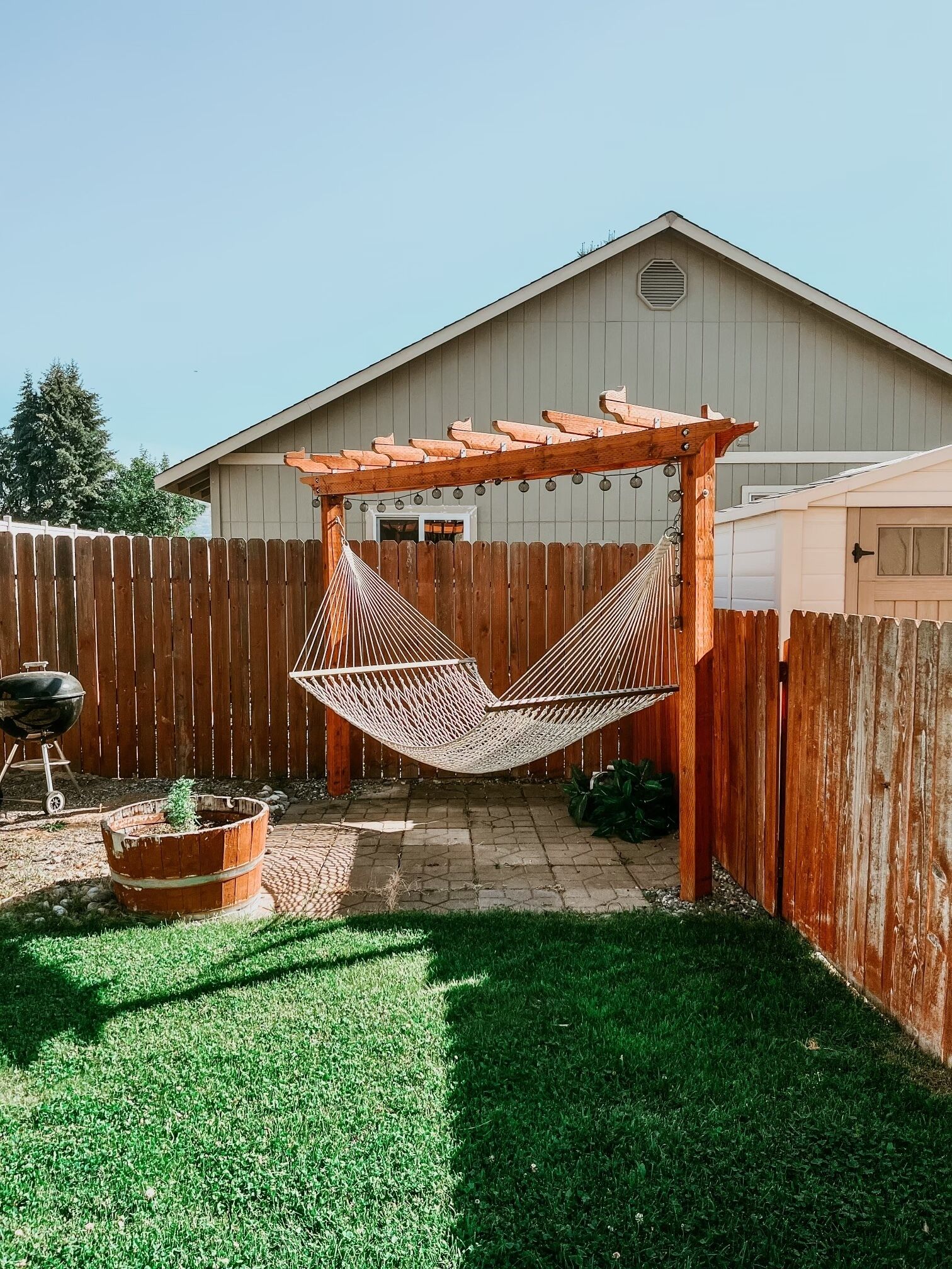 A hammock under a wooden pergola in a backyard with a wooden fence, a barbecue grill, and a potted plant.