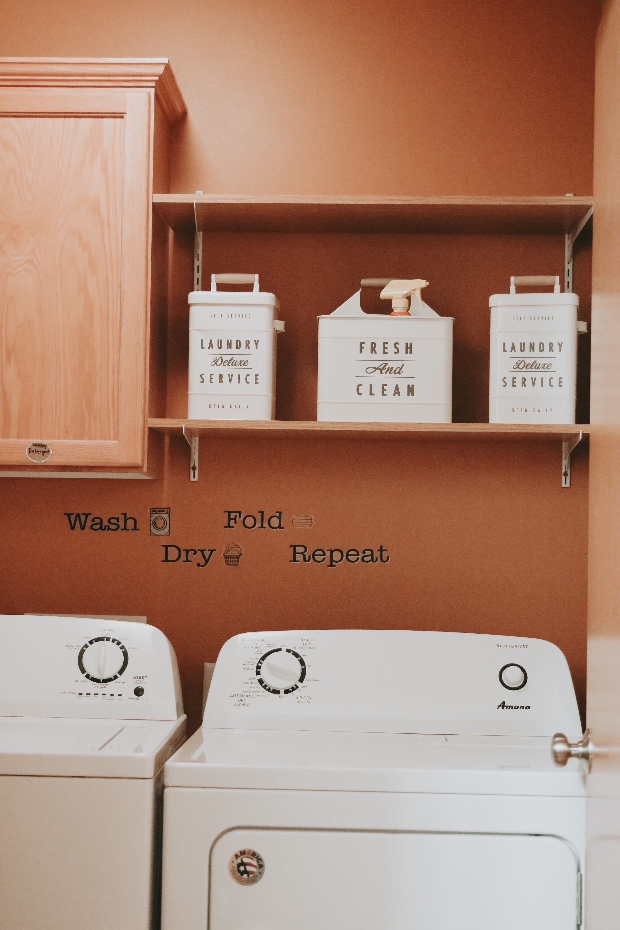 A laundry area with two white washing machines, wooden cabinets, and shelves with detergent bottles labeled 'Laundry Deluxe Service' and 'Fresh and Clean'.