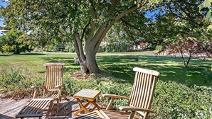 A wooden deck with three Adirondack chairs and a small table, surrounded by lush greenery and a large tree. - 4 Unique Cottages Sleeps 16 Comfortably on 1 Acre (Coloma)