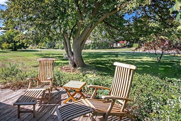 Una terraza de madera con tres sillas Adirondack y una mesita pequeña, rodeada de abundante follaje y un árbol grande.