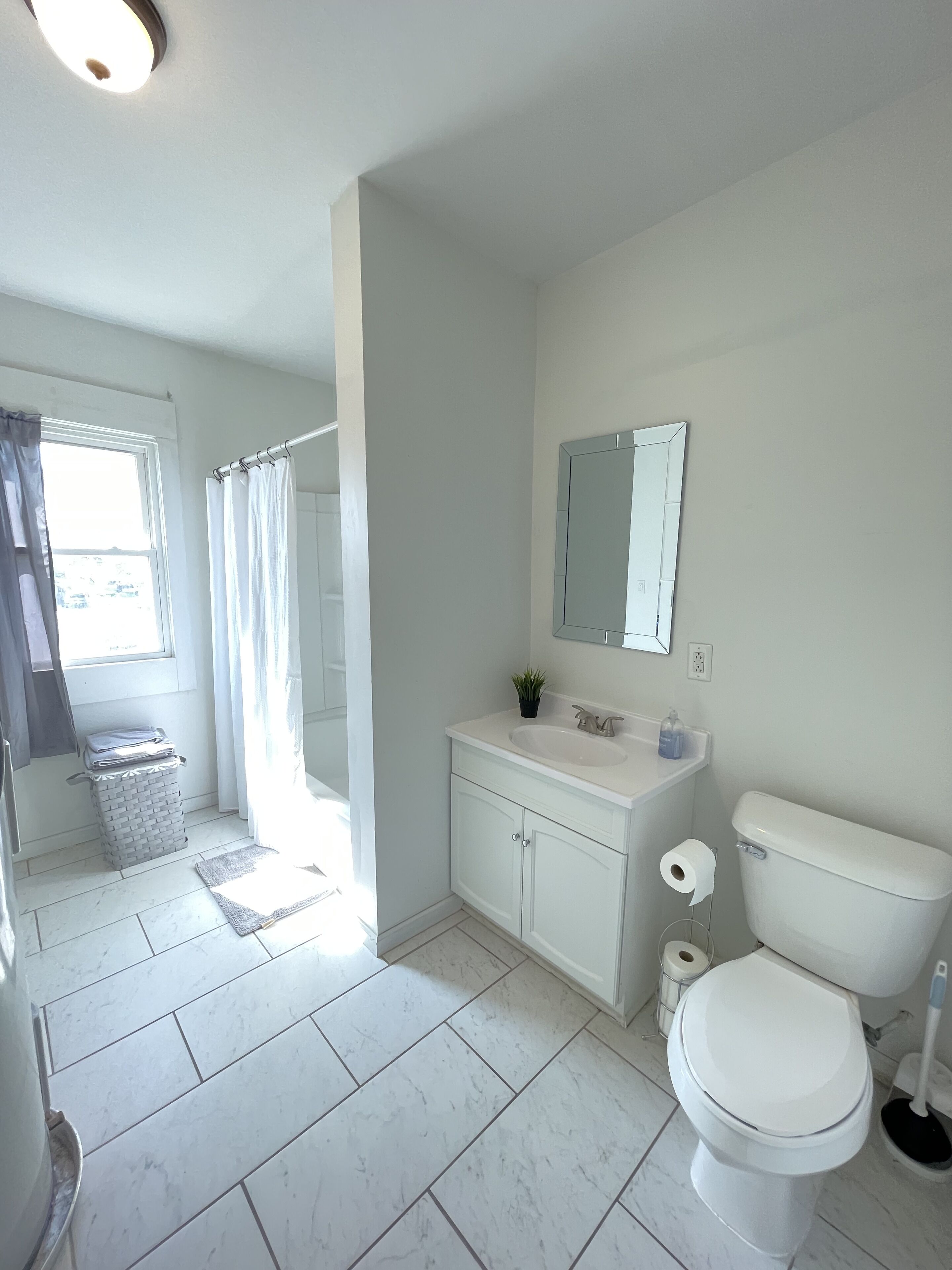 Une salle de bain avec des toilettes, un lavabo et un miroir. Il y a une fenêtre avec des rideaux et une petite poubelle.