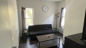 Quarto com ventoinha no teto, sofá preto, mesa de centro, relógio e janela com cortinas.