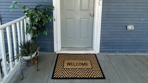 Uma porta de entrada com um tapete de boas-vindas, um vaso de planta e um corrimão branco.