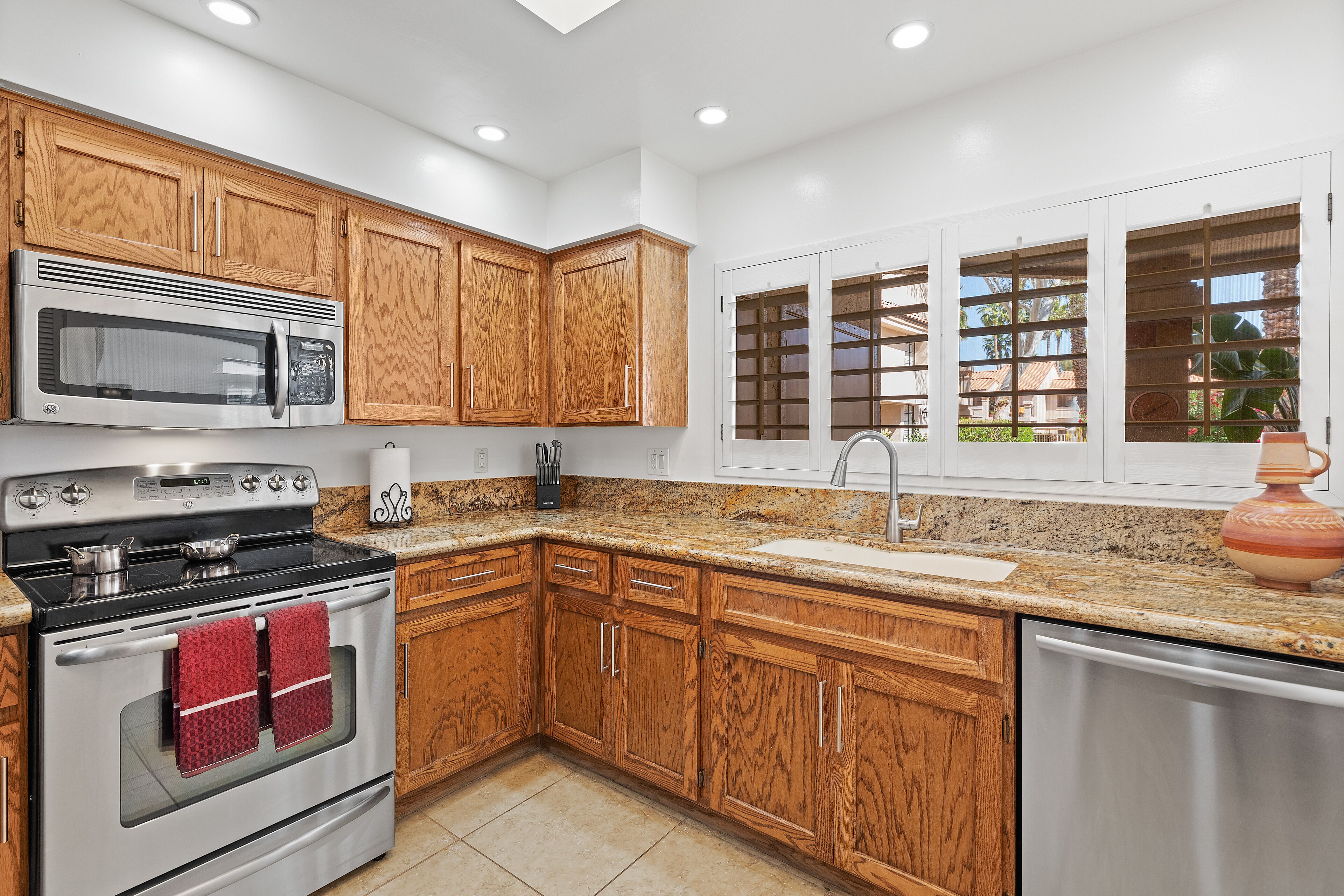 A kitchen with wooden cabinets, stainless steel appliances, a granite countertop, and a window with a view of greenery.