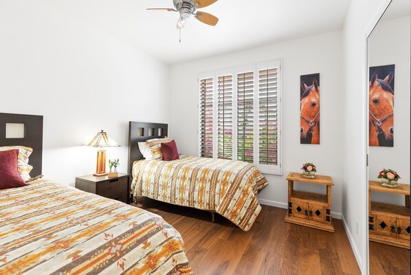 A bedroom with two beds, a ceiling fan, a window with shutters, and a small wooden nightstand with a vase.