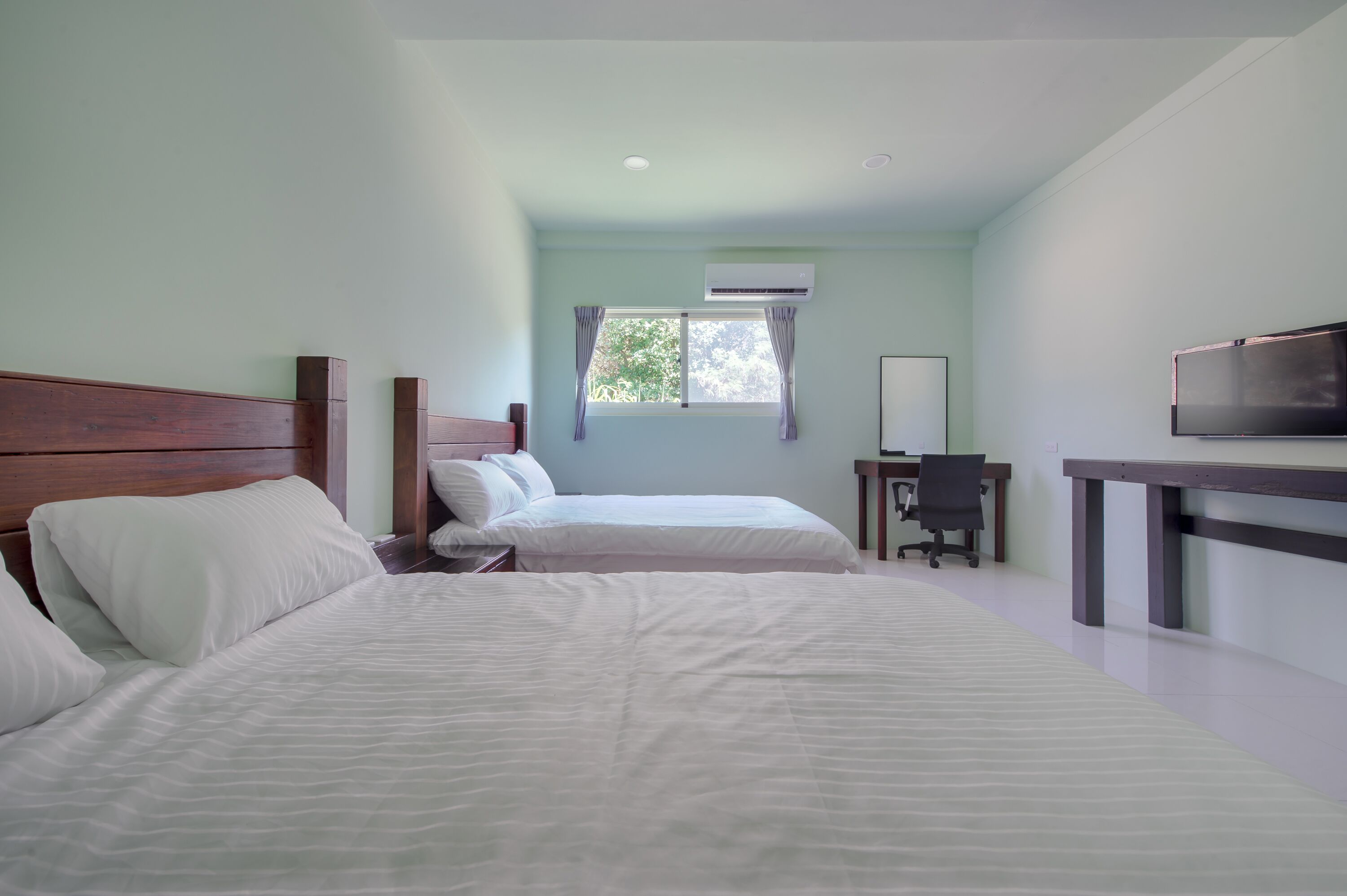 A hotel room with two beds, a desk, a chair, a TV, and an air conditioning unit.