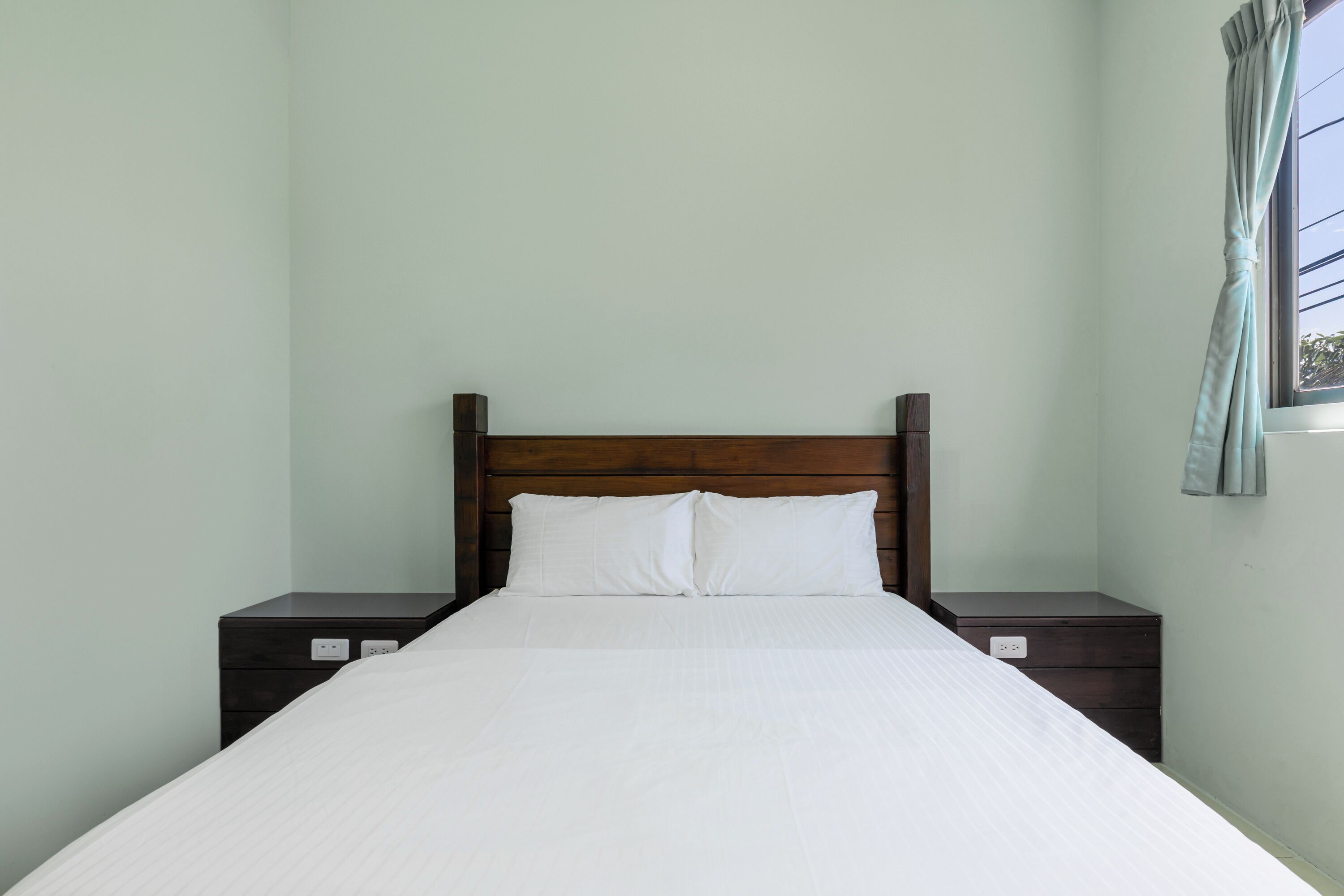 A neatly made bed with a wooden headboard, two bedside tables, and a window with curtains.