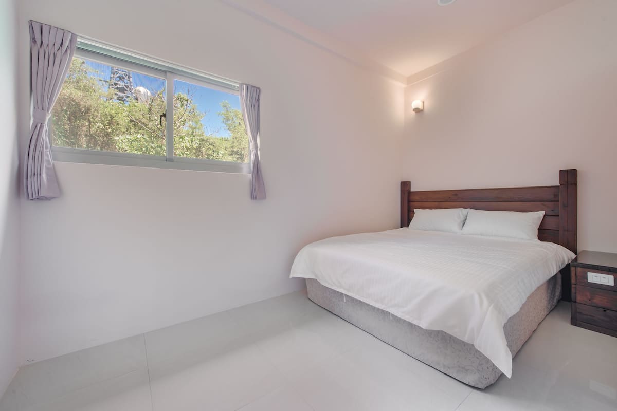 A bedroom with a bed, a nightstand, a window with curtains, and a wall-mounted light.